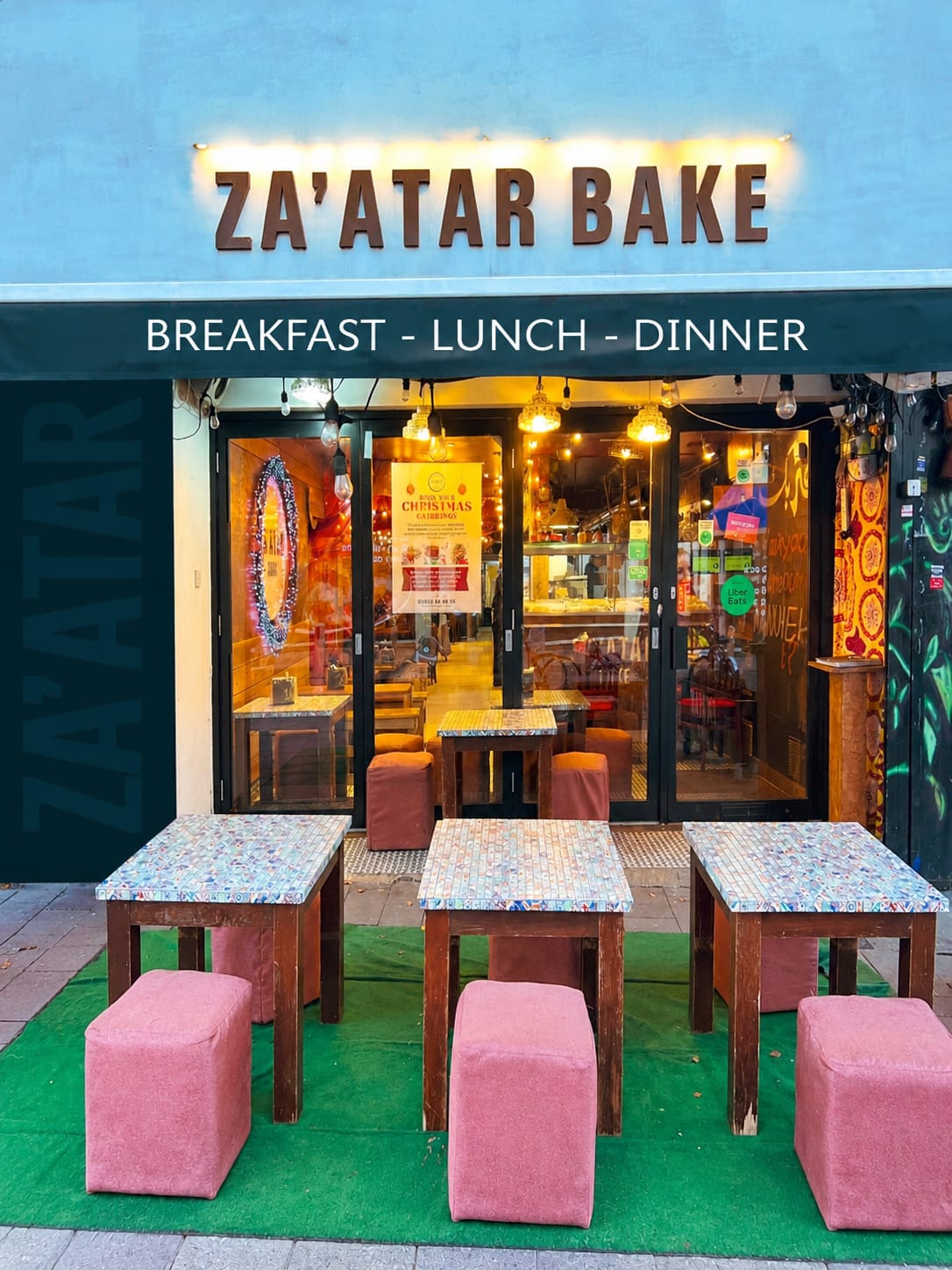 Za'atar Bake storefront showing illuminated restaurant signage and outdoor seating with tiled tables and pink ottomans